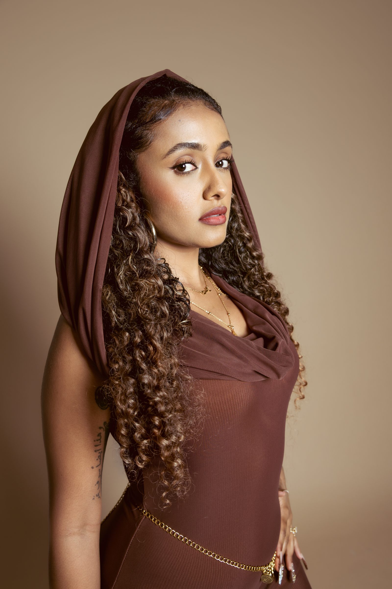 Sharin on a sand backdrop, her cocoa headscarf drawn up over long curls, looking into the camera, a gold coin belt at her waist