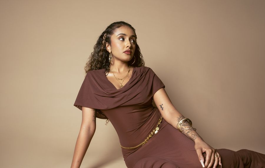 Sharin seated on a sand backdrop in a cocoa dress and gold coin belt, looking up and to the side